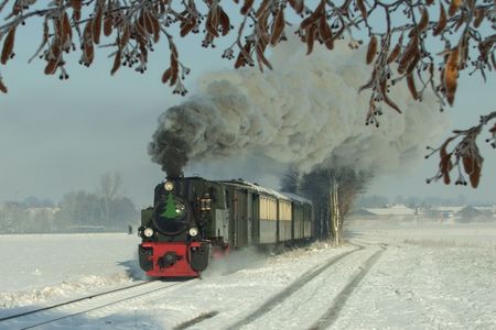 Eine historische, dampfende Lok mit mehreren Waggons fährt auf einem Gleis durch eine Schneelandschaft.