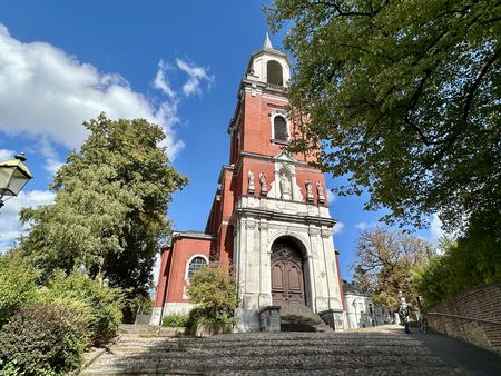 Bäume umrahmen eine Kirche aus rötlichem und weißem Stein, zu der Treppen hinaufführen.