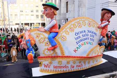 Eine kunstvoll gestalteter Karnevalswagen mit zwei Figuren fährt vor einer Reihe Zuschauenden auf der Straße.