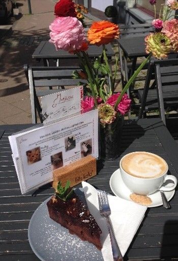 Bei sonnigem Wetter steht ein Kaffee und ein Stück Kuchen auf einem Tisch, zusammen mit Blumen und einer Speisekarte.