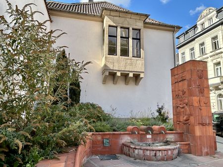 Rötlicher Brunnen mit drei Quellen vor eine weißen Haus.