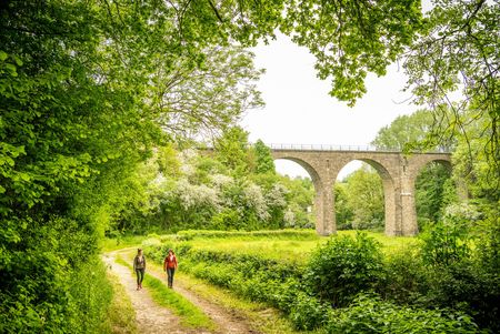 Zwei Personen wandern auf einem Feldweg inmitten von saftig grünen Wiesen und Bäumen und einer Steinbrücke mit Bögen im Hintergrund.
