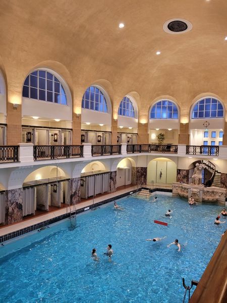 Schwimmhalle mit großem Schwimmbecken, Wandleuchten, hoher gewölbter Decke und Bogenfenstern. 