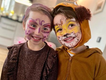 Zwei verkleidete und geschminkte Kinder stehen eng nebeneinander und lachen, eines ist als Löwe und das andere als Schmetterling verkleidet.