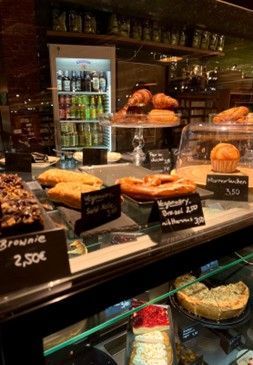 Auf einer Theke steht verschiedenes Gebäck mit Namens- und Preisschildern. Im Hintergrund steht ein Getränkekühlschrank.