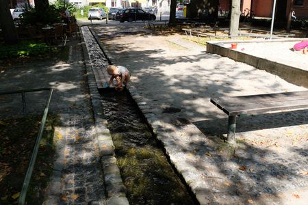 An einem kleinen Bach entlang einer Straße spielt ein Kind. Rechts befindet sich ein Sandkasten.