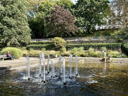 Springbrunnen mit mehreren Fontänen inmitten eines teilweise gepflasterten Parks mit Bäumen.