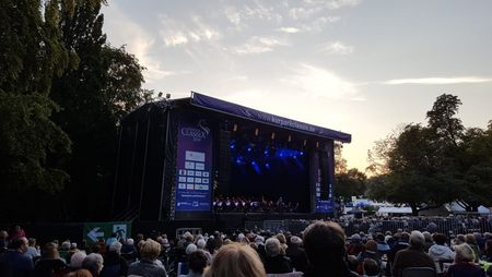 Unter freiem Abendhimmel ist eine Bühne aufgebaut, auf der ein Konzert stattfindet und viele Besuchende davor sitzen.
