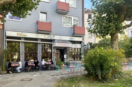 Außenansicht eines Restaurants, vor dem mehrere Personen an Tischen speisen, umrahmt von Bäumen.